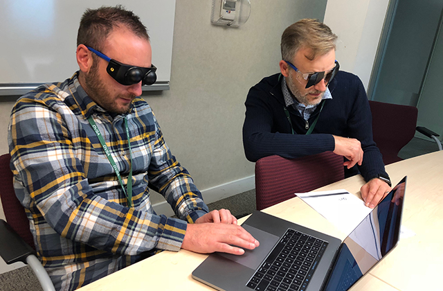 Image of one man wearing visual impairment simulation googles using a laptop, and another man wearing visual impairment simulation goggles looking at a sheet of paper